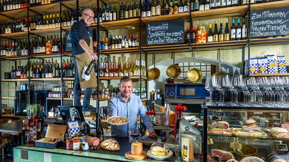 Due uomini in un ristorante con cibi e bevande, uno serve e l'altro prepara un piatto. | © Paul Stajan