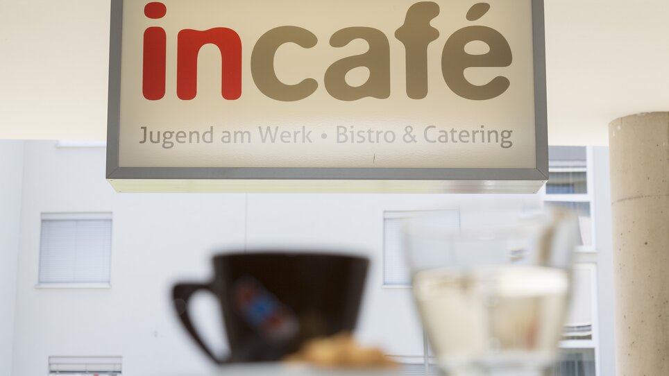 Insegna con la scritta “incafé - Jugend am Werk - Bistro & Catering”. In primo piano si vedono una tazza di caffè e un bicchiere d'acqua. | © incafe