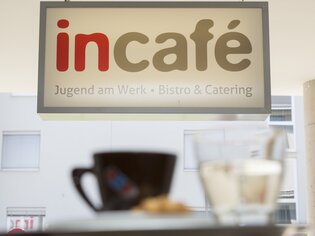 Insegna con la scritta “incafé - Jugend am Werk - Bistro & Catering”. In primo piano si vedono una tazza di caffè e un bicchiere d'acqua. | © incafe