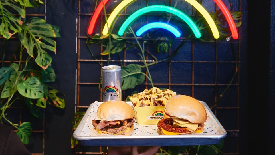 Zwei Burger, Pommes und eine Getränkedose vor einer Neo-Lampe in Form eines Regenbogens. | © Samuel Kuro