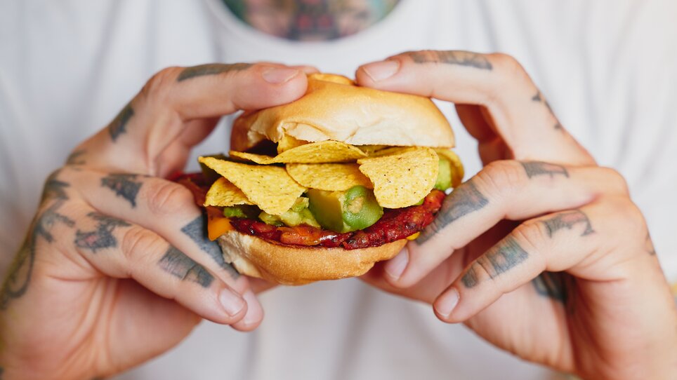 Ein Mann hält einen Hamburger mit Nachos und Avocado-Stücken in seinen Händen. | © Samuel Kuro