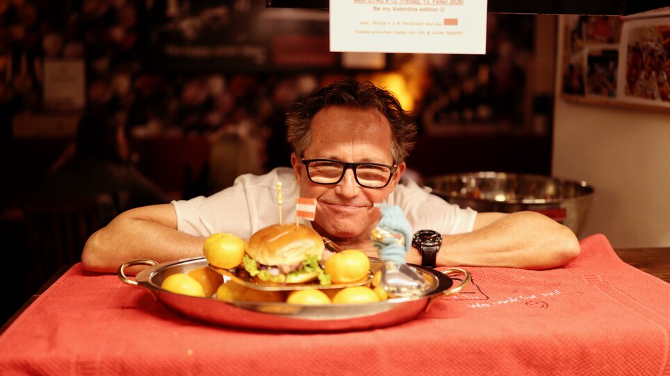The manager of Kitchen12 smiles behind a tray of burgers and potatoes. | © Makinson Media