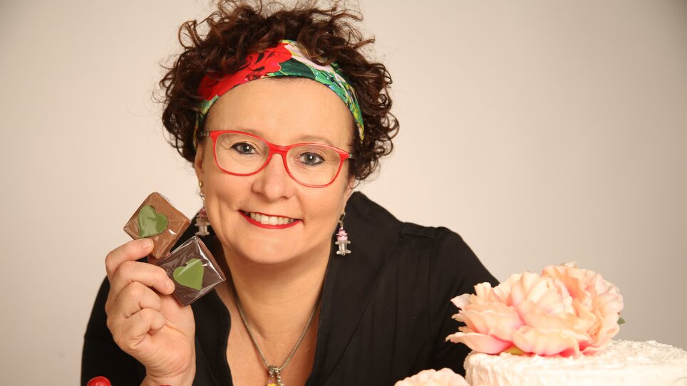A woman with red glasses holds chocolates in front of cakes and desserts.