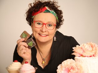 Una donna con occhiali rossi tiene cioccolatini davanti a torte e dolci.