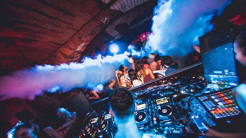 A DJ performs in a club surrounded by fog and dancing guests.