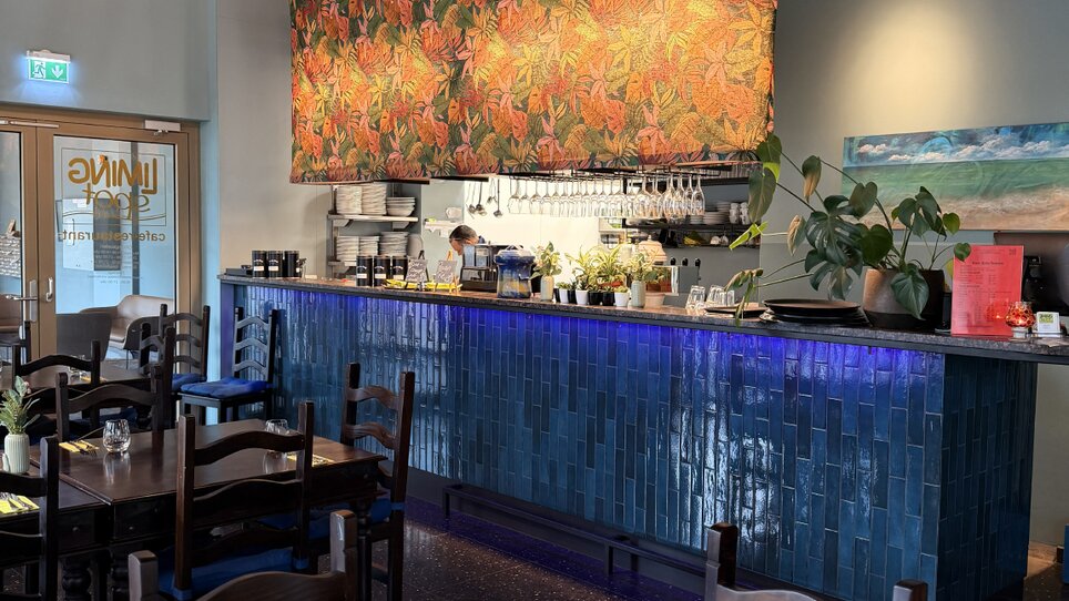 Modern restaurant interior with colorful wall hanging and blue bar counter. | © Foto Vision