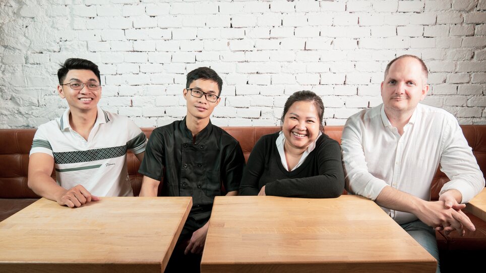 Four people sitting at a table in a restaurant with a brick wall. | © Ma Kitchen