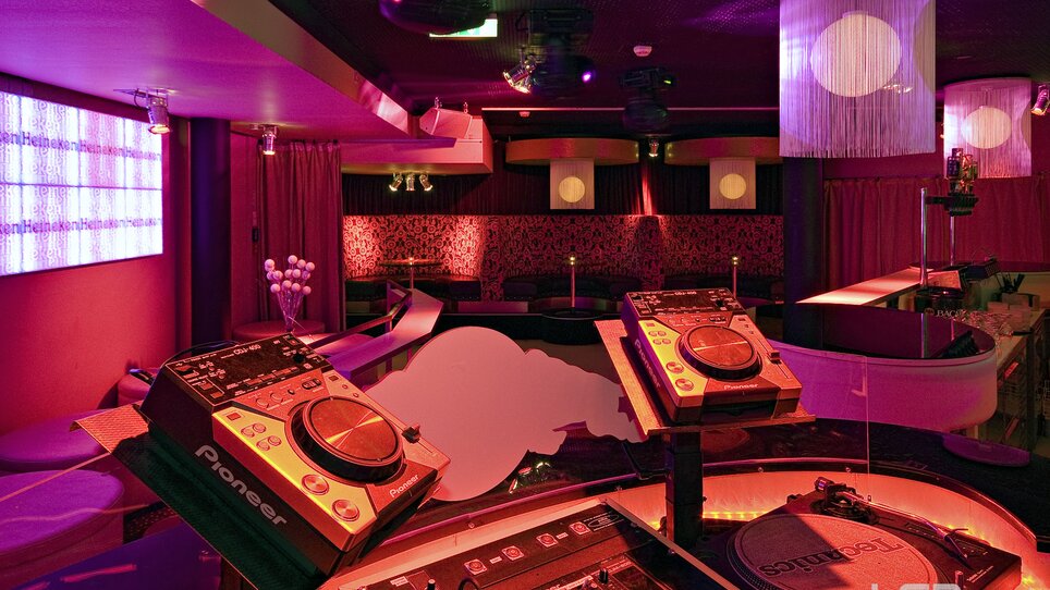 Vista di un club elegante con attrezzatura DJ e luci colorate. | © Mausefalle +