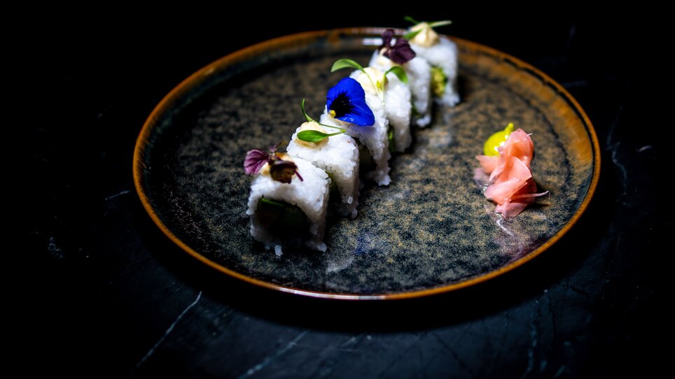 Sushi artistico con fiori commestibili e zenzero su un piatto nero.
