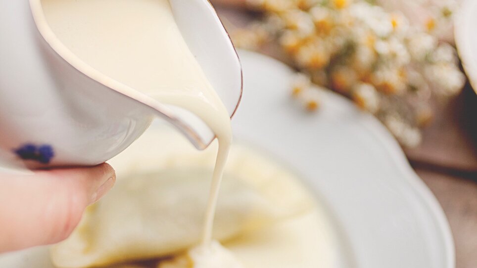 A hand pouring creamy sauce over pierogi on a plate.