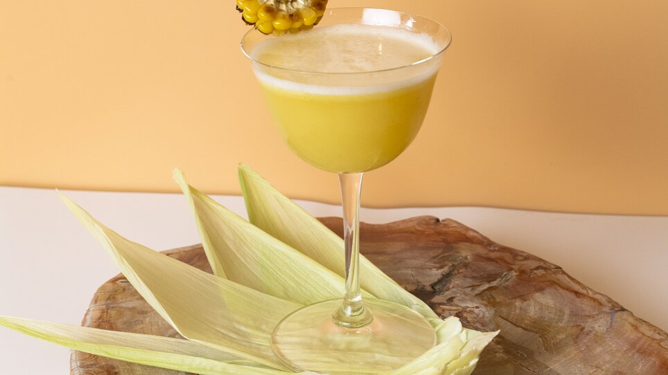 A yellow cocktail in an elegant glass, garnished with a slice of corn. | © Jauk Katharina