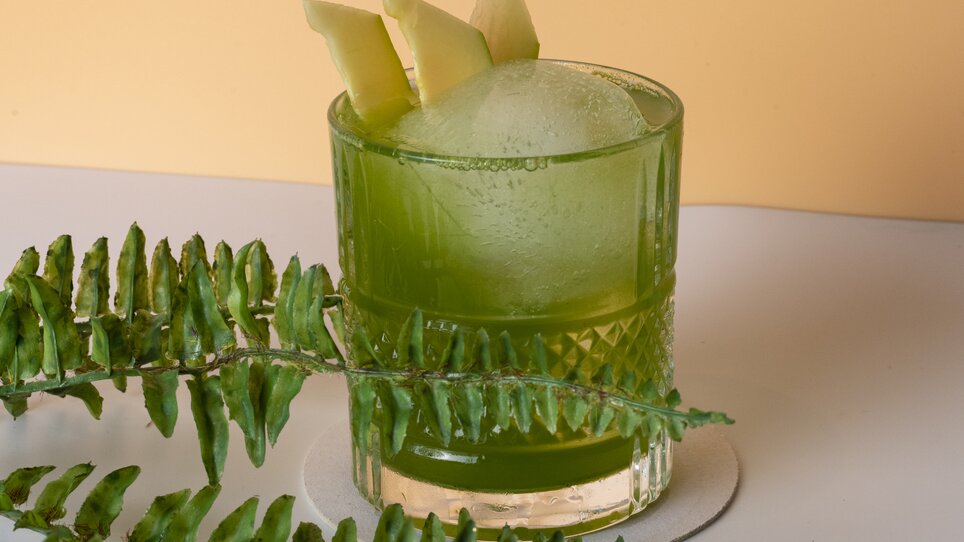 A green cocktail with apple slices, surrounded by ferns. | © Jauk Katharina