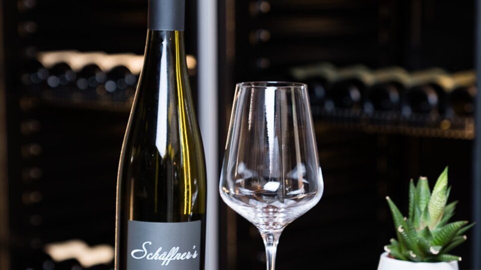 Ein Weinflasche und ein Glas auf einem Tisch, im Hintergrund ein Weinregal. | © Schäffner