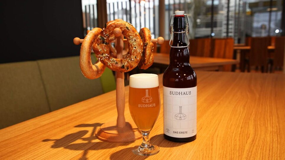 Ein Glas Bier und eine Flasche Sudhaus Bier neben einem Ständer mit Brezeln auf einem Tisch.