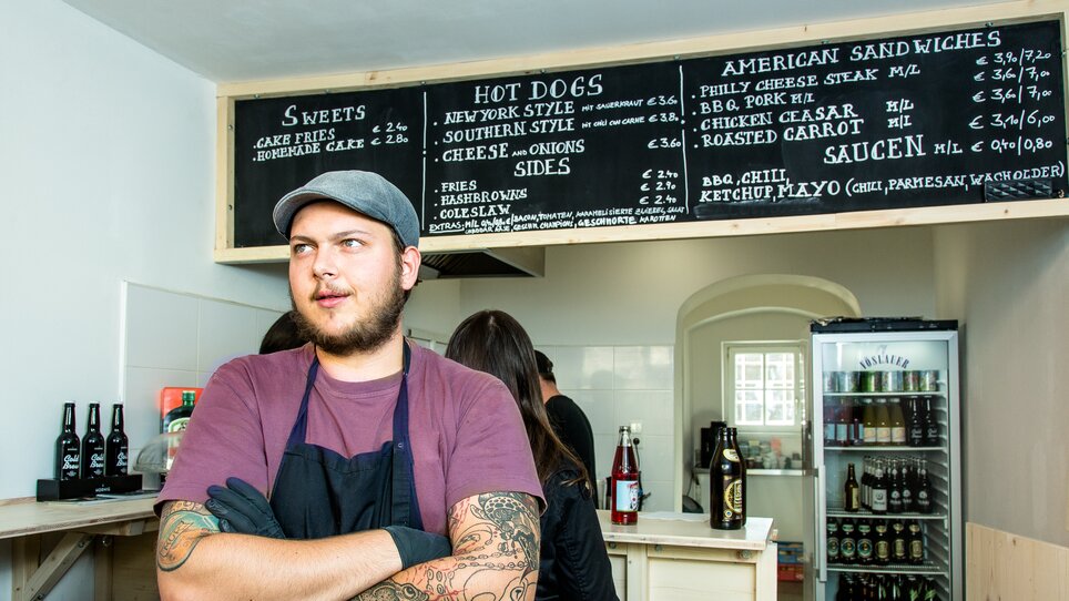 Man behind the counter of a restaurant with a menu above. | © The Hungry Heart