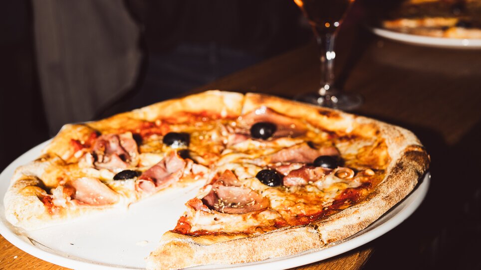 Ein Teller Pizza mit Oliven und Schinken, dazu ein Drink auf einem Tisch in einer Bar. | © Chiara Maria B.