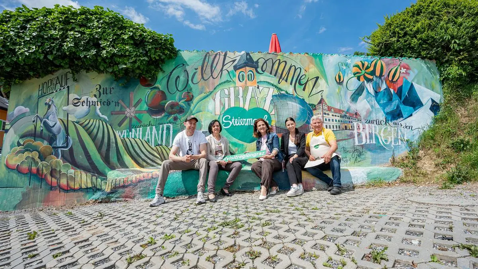 Fünf Personen posieren vor einem bunten Wandbild - gestaltet von Tom Lohner - in Graz. | © STG - Jaden Gynes