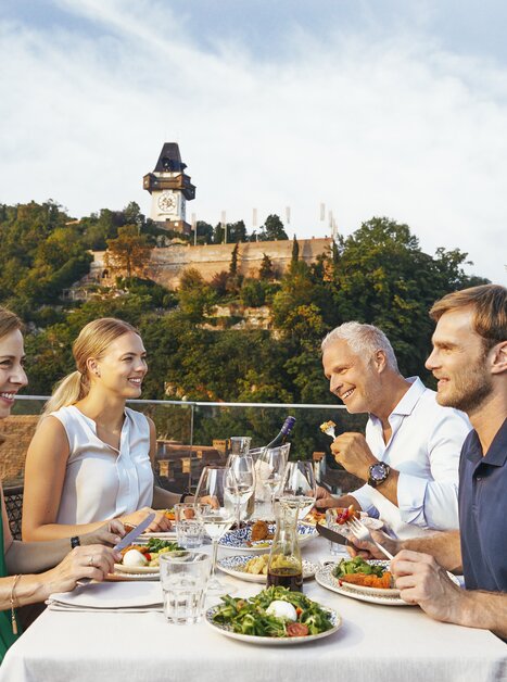 Vier Personen genießen ein festliches Essen auf einer Terrasse mit Blick auf den Grazer Uhrturm. | © Graz Tourismus - Astrid Schwab