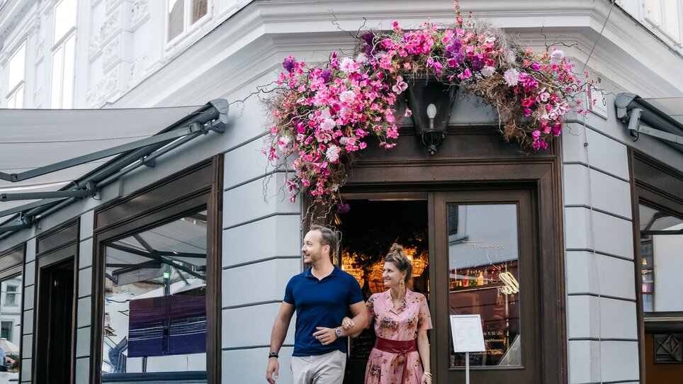 Una coppia esce da un ristorante a Graz, circondata da piante in fiore. | © Graz Tourismus - Mias Photoart-48