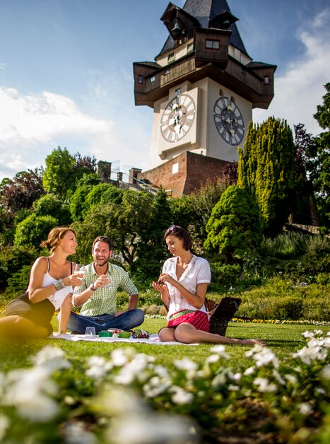 Freunde genießen eine Auszeit am Schlossberg in Graz, mit Grazer Uhrturm im Hintergrund und picknicken gemeinsam. | © Graz Tourismus - Tom Lamm