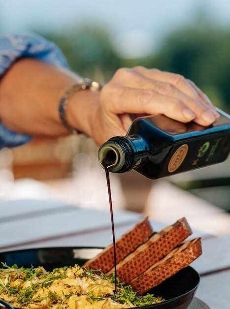 Eine Hand gießt Kernöl aus einer Glasflasche über eine Eierspeise mit Kräutern und Brot. | © Graz Tourismus_Mias Photoart