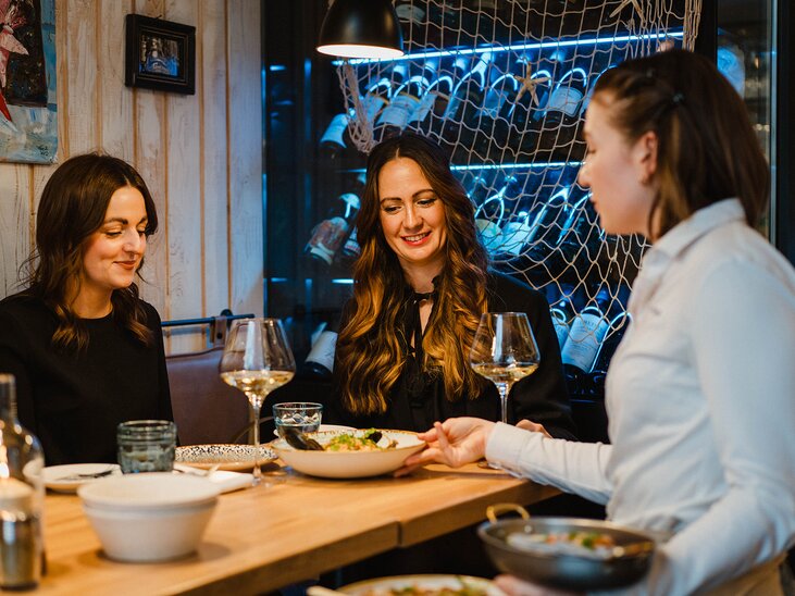 Zwei Frauen genießen ihr Essen in einem Restaurant, eine Kellnerin serviert die Gerichte. | © 5komma5sinne- MotionAds