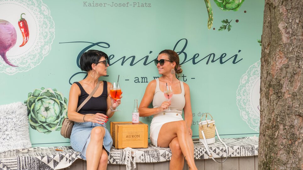 Due donne gustano drink a Graz davanti a un muro con scritte. | © 5komma5sinne - Helmut Schweighofer