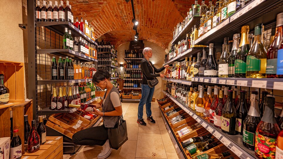 Persone che acquistano in un negozio di vini con scaffali pieni di bottiglie. | © 5komma5sinne - Rene Strasser Fotografie