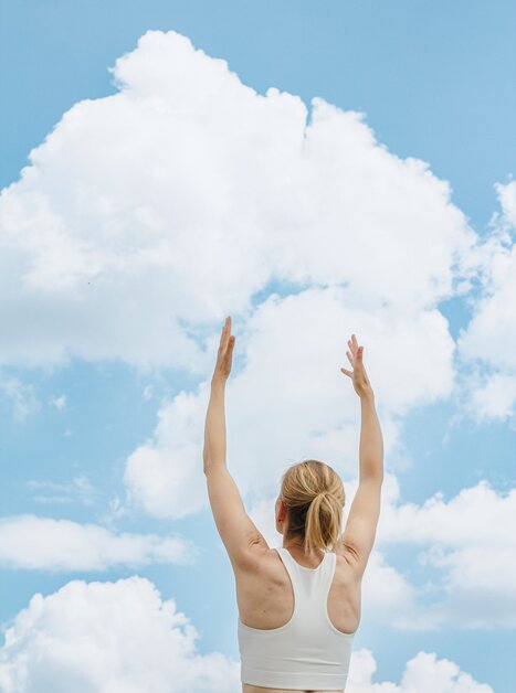 A blonde woman in sportswear stretches both arms upwards, a blue sky with clouds can be seen behind her | © Augartenhotel x einsnullneun