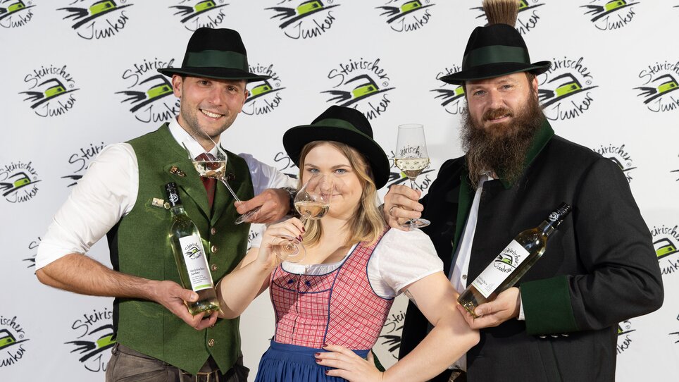 Drei elegante Personen in Tracht mit Weinflaschen und Gläsern in den Händen posieren vor dem Hintergrund einer Logo-Wand mit dem Logo des Steirischen Junkers. | © Wein Steiermark  - Fotokuchl