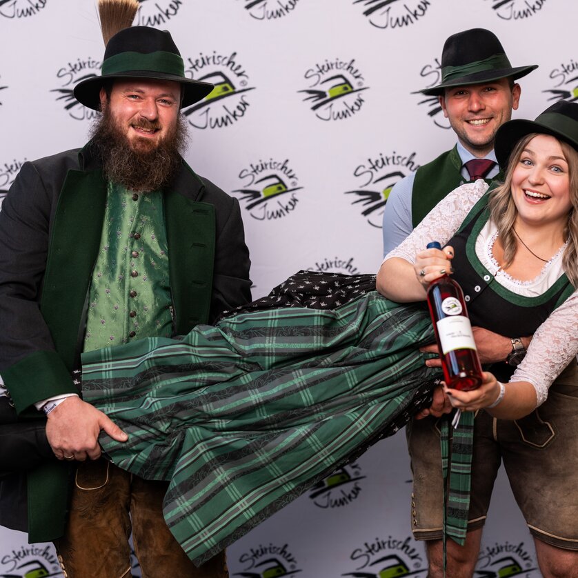 Eine fröhliche Gruppe in Tracht präsentiert eine Flasche Wein auf einer Veranstaltung. | © Wein Steiermark - Fotokuchl
