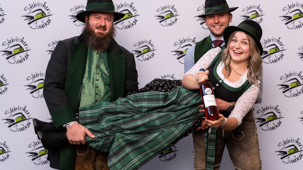 Eine fröhliche Gruppe in Tracht präsentiert eine Flasche Wein auf einer Veranstaltung. | © Wein Steiermark - Fotokuchl