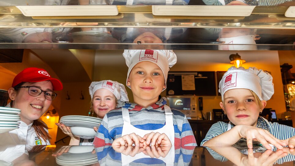 Drei Kinder mit Kochmützen und eine Köchin lächeln bei einem Kochkurs in die Kamera. | © Graz Tourismus - Paul Stajan