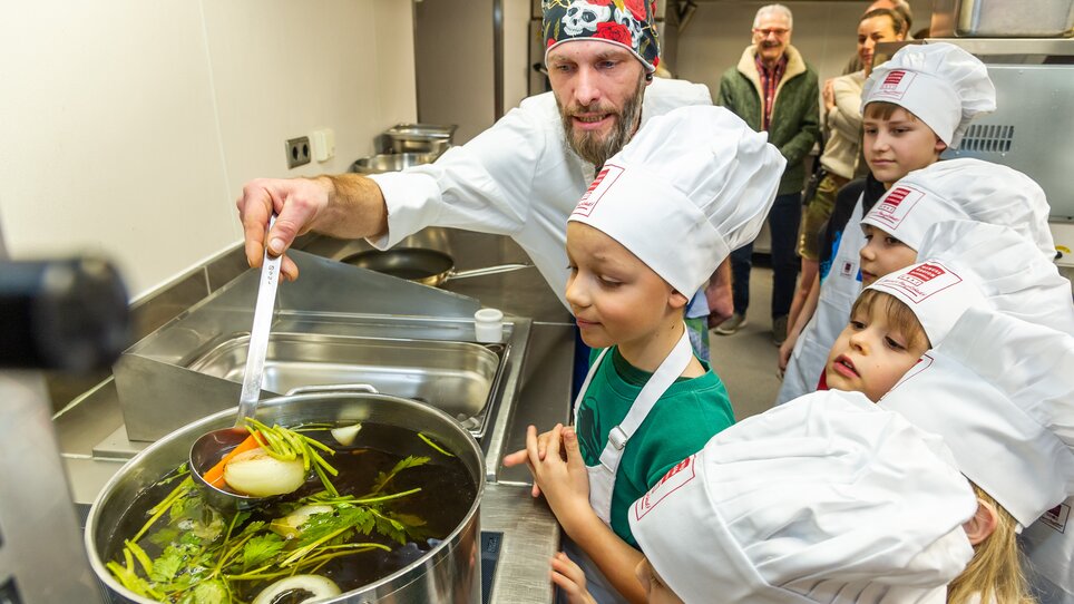 Ein Koch zeigt Kindern bei einem Kochkurs, wie sie Gemüse in einen Topf geben. | © Graz Tourismus - Paul Stajan