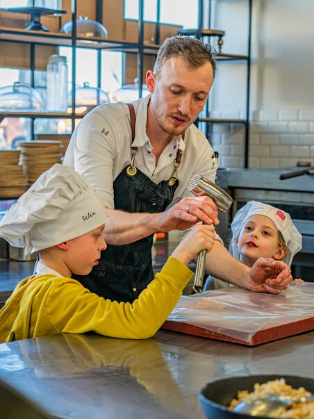 Ein Koch in einer Küche zeigt Kindern bei einem Kochkurs, wie man Teig zubereitet. | © Milena Bonstingl
