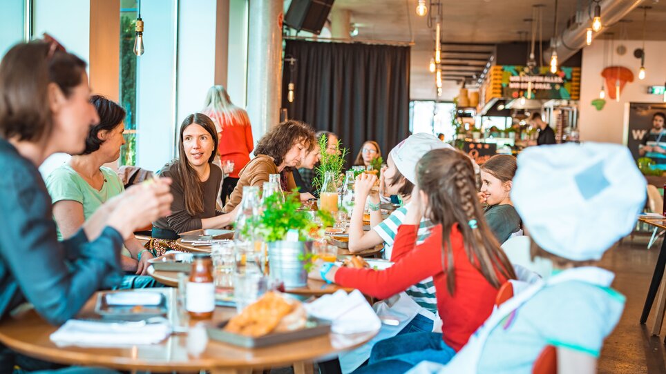 Eine Gruppe von Kindern und Erwachsenen sitzt an einem Tisch, um das beim Kinder-Kochkurs zubereitete Essen zu genießen. | © KHC - Bernhard Schindler