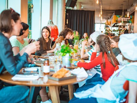 Eine Gruppe von Kindern und Erwachsenen sitzt an einem Tisch, um das beim Kinder-Kochkurs zubereitete Essen zu genießen. | © KHC - Bernhard Schindler