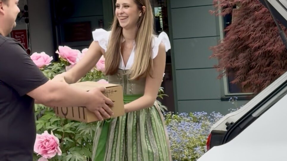 Donna in abiti tradizionali consegna una scatola a un uomo, circondata da fiori. | © Schanzlwirt