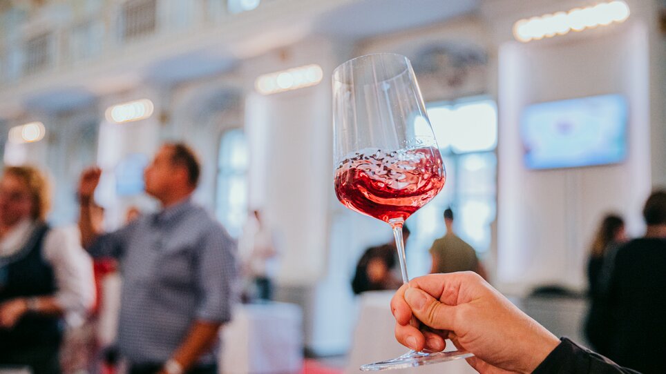 Ein Weinglas mit rot schimmerndem Wein wird in die Kamera gehalten. Der Wein wirbelt im Glas, im Hintergrund sind unscharf Menschen zu erkennen. Die Atmosphäre wirkt festlich und stilvoll. | © Johanna Lamprecht