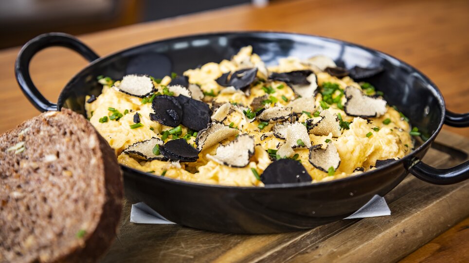 A dish featuring egg, black truffles, and fresh herbs in a black bowl, alongside a piece of bread. | © Graz Tourismus - Werner Krug