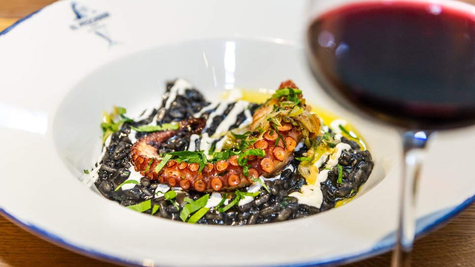 Ein Teller mit schwarzem Risotto, Octopus und Garnitur, begleitet von einem Glas Rotwein. | © Paul Stajan