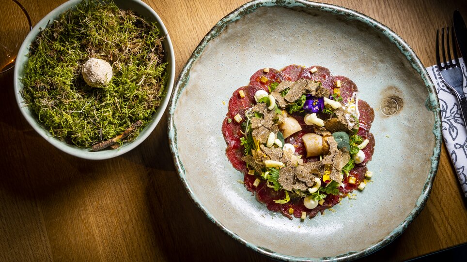 An elegant dish with slices of meat, truffles, and decoration on a plate, next to a bowl with moss. | © Graz Tourismus - Werner Krug