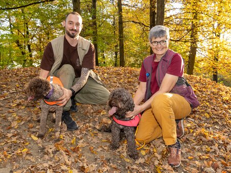 A man and a woman pose with two dogs in the woods during autumn. | © Graz Tourismus - Harry Schiffer