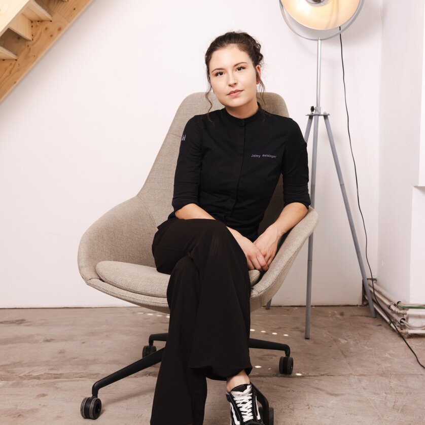 Chef-Patissière Jaimy Reisinger in schwarzem Outfit sitzt entspannt auf einem grauen Stuhl. | © Pixelcoma