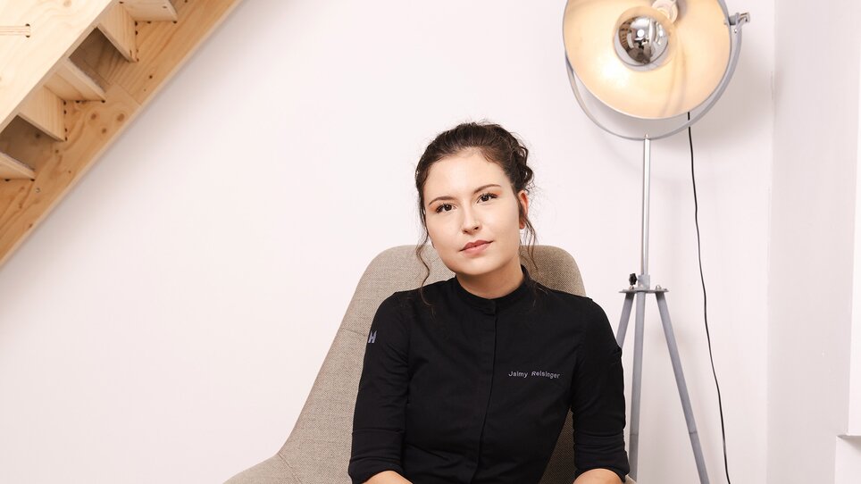 La chef pasticcera Jaimy Reisinger, vestita di nero, è seduta rilassata su una sedia grigia. | © Pixelcoma