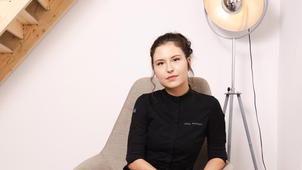 Chef-Patissière Jaimy Reisinger in schwarzem Outfit sitzt entspannt auf einem grauen Stuhl. | © Pixelcoma
