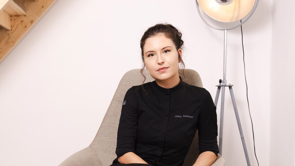 Chef-Patissière Jaimy Reisinger in schwarzem Outfit sitzt entspannt auf einem grauen Stuhl. | © Pixelcoma