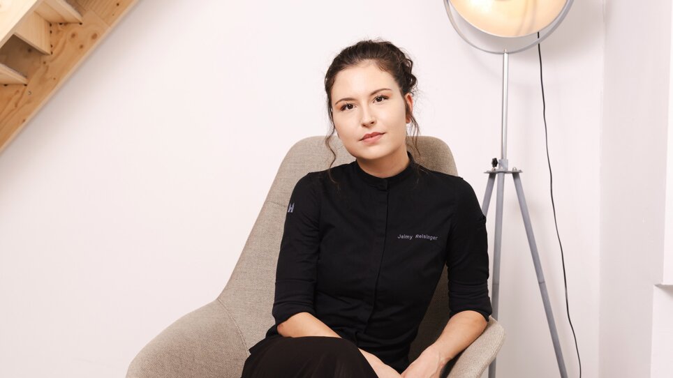 Chef-Patissière Jaimy Reisinger in schwarzem Outfit sitzt entspannt auf einem grauen Stuhl. | © Pixelcoma