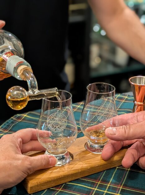 Zwei Gläser werden mit Whiskey gefüllt, während zwei Hände bereit sind. | © The Islay Whisky Chapter Austria
