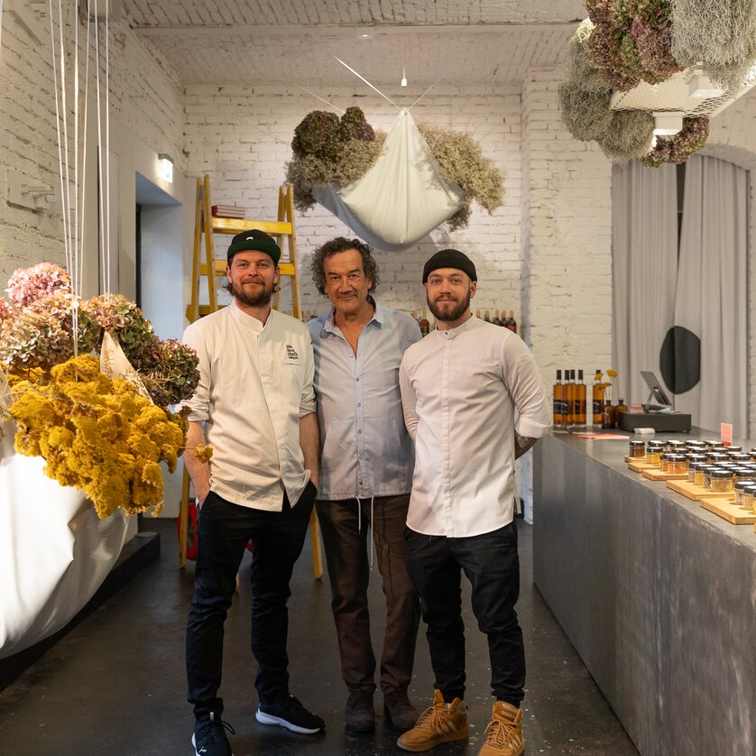 Drei Köche stehen in einem Gastronomieladen mit Trockenblumen und Produkten. | © Michael Kink